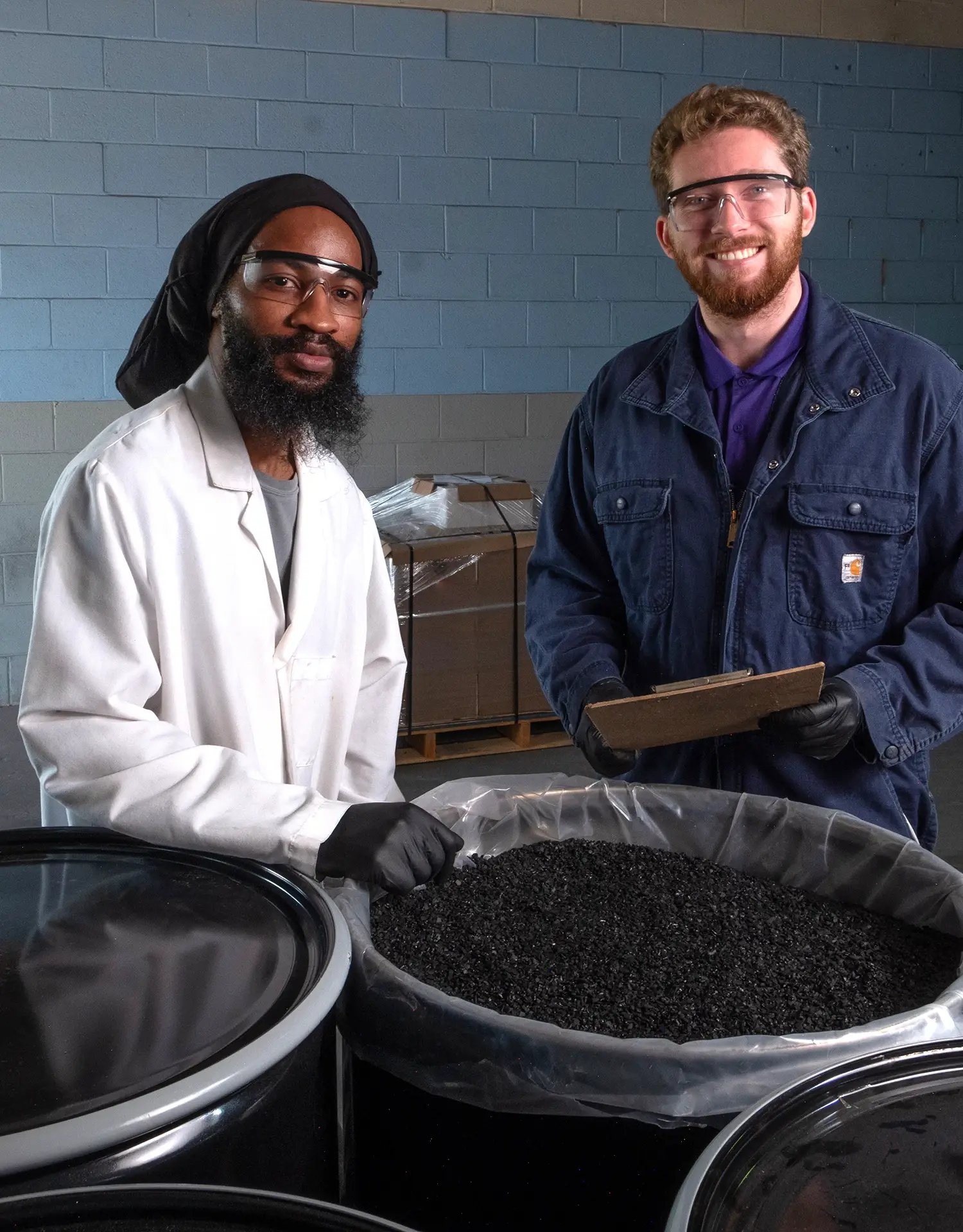 Two ActiveSite team members wearing safety glasses stand beside a large container of black catalyst material in a manufacturing facility.